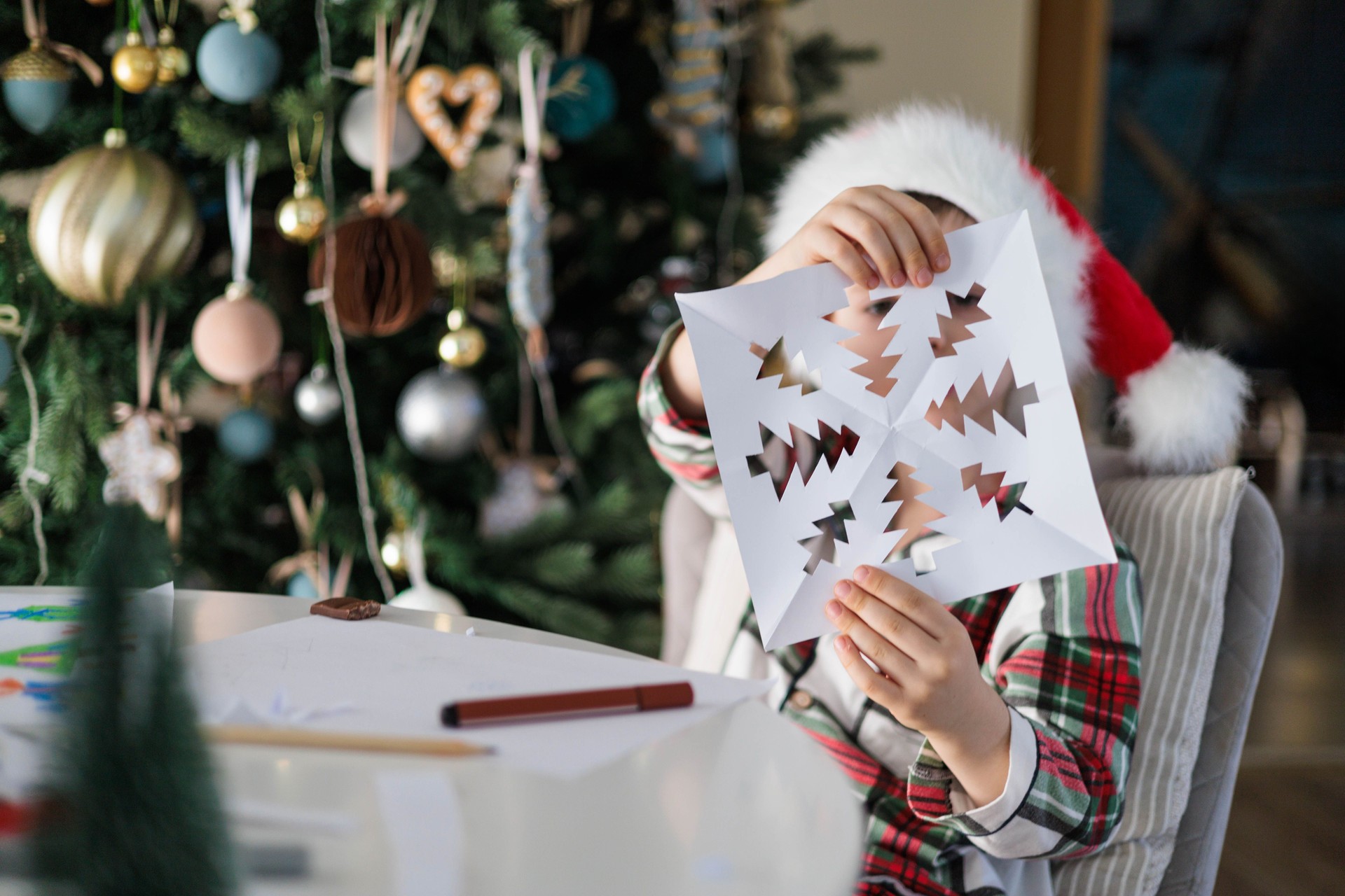 Child Showing Handmade Christmas Paper Craft in Santa Hat near Festive Tree at Home