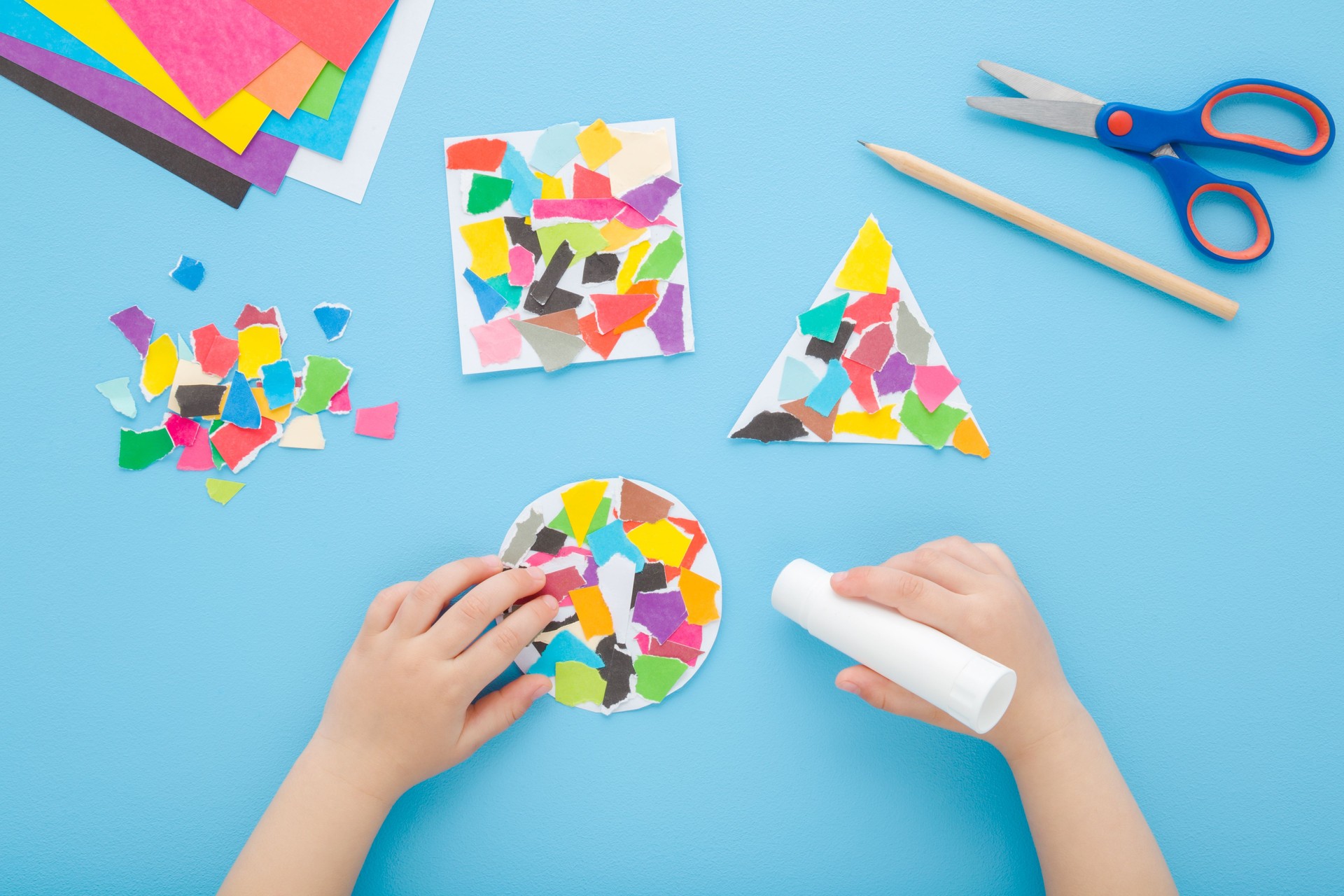 Baby boy hands gluing different geometric shapes with colorful torn paper pieces. Blue table background. Pastel color. Closeup. Development task for little kid motor skills. Top down view.